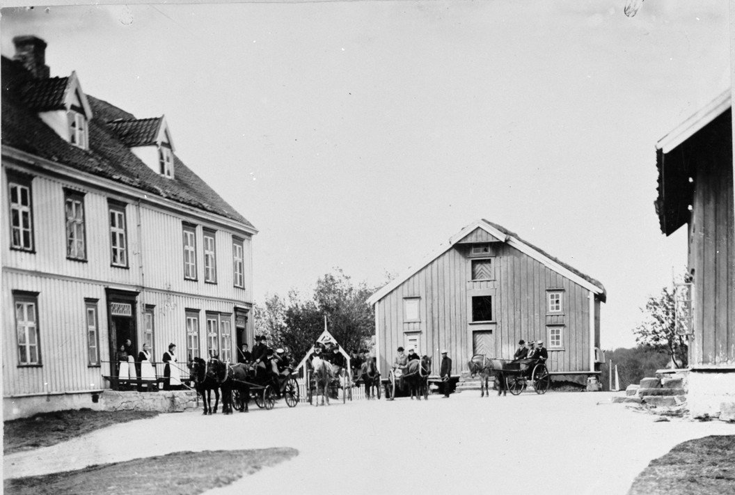 Tun mellom tre historiske bygninger. Til venstre i bildet et større trehus i empirestil. Midt i front et stort stabbur med torvtak. På gårdsplassen er der mange mennsker, både kvinner i lange kjoler, arbeidskarer, tjenestejenter og andre. På gårdsplassen er der også to vogner med hester spent for.